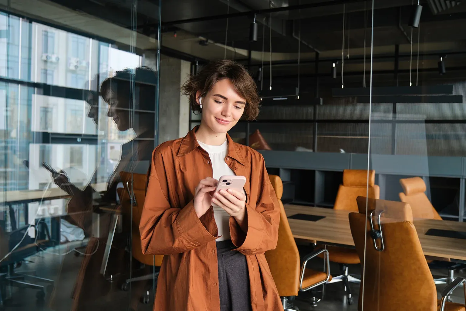 Mujer joven y sonriente mirando su celular en una oficina moderna y cálida. Representa la facilidad de gestionar las finanzas de una empresa en tiempo real desde el teléfono o WhatsApp.