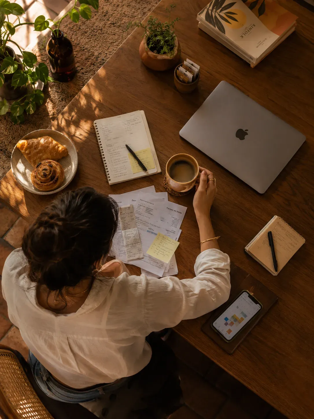 Vista superior de una mujer organizando facturas y gastos de su negocio en una mesa de madera con su computador y un café.
